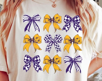 Può includere: Una maglietta bianca con un design di nove fiocchi gialli e viola. I fiocchi sono disposti in uno schema a griglia 3x3.