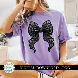 May include: A purple t-shirt with a large black bow design. The bow is made of a sparkly material and has a 3D effect. The shirt is a casual style and has a relaxed fit.