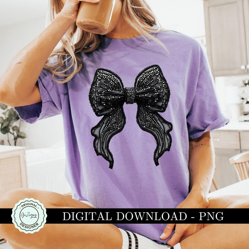 May include: A purple t-shirt with a large black bow design. The bow is made of a sparkly material and has a 3D effect. The shirt is a casual style and has a relaxed fit.
