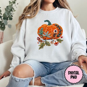 Peut inclure: Un sweat-shirt blanc avec un motif brod&eacute; d'une citrouille orange avec des feuilles vertes et des fleurs. La citrouille est entour&eacute;e de feuilles vertes et de petites fleurs dans des tons de rose, orange et blanc.