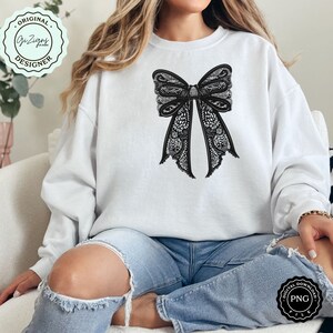 May include: A white sweatshirt with a black lace bow design on the front. The sweatshirt is being worn by a person sitting on a white chair.