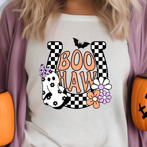 May include: A cream-colored t-shirt featuring a Halloween-themed design. The design includes the words "BOO HAW" in orange, a ghost, flowers, a bat, and a horseshoe with a black and white checkered pattern.