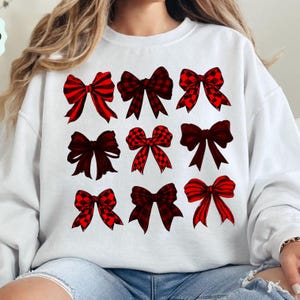 May include: White sweatshirt featuring nine red bows with various patterns, including plaid and solid designs. The bows are arranged in three rows of three, offering a festive and decorative aesthetic. The bows are in shades of red and black.