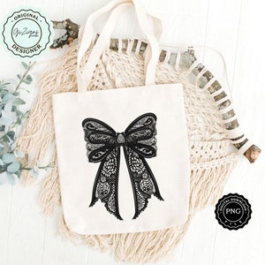 May include: A white tote bag with a black lace bow design. The bag is on a white wooden surface with a macrame wall hanging in the background.