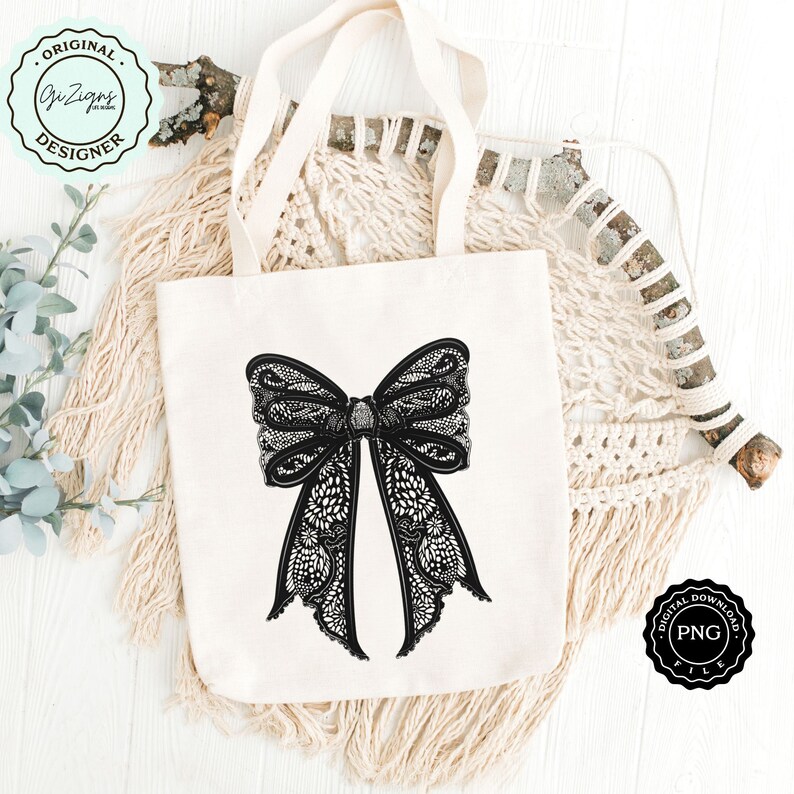 May include: A white tote bag with a black lace bow design. The bag is on a white wooden surface with a macrame wall hanging in the background.