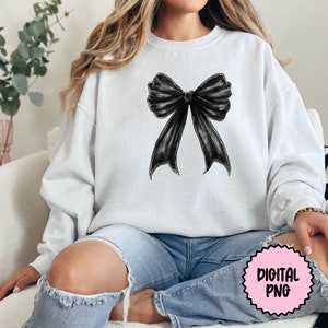 Pode incluir: Uma camisa de moletom branca com um grande la&ccedil;o preto estampado na frente. A camisa de moletom est&aacute; sendo usada por uma pessoa sentada em um sof&aacute; branco.