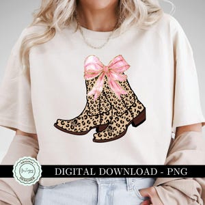 K&ouml;nnte beinhalten: Ein wei&szlig;es T-Shirt mit dem Text "Digitaler Download - PNG" am unteren Rand. Das Shirt zeigt ein Paar braune und schwarze Cowboy-Stiefel im Leopardenmuster mit einer rosa Schleife oben.