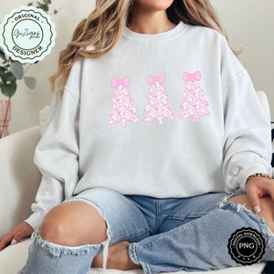 Pu&ograve; includere: Una felpa bianca con un design rosa di tre alberi di Natale. Ogni albero &egrave; composto da piccoli fiocchi rosa e ha un fiocco rosa pi&ugrave; grande in cima.