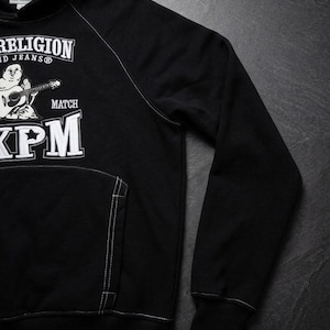 Op de afbeelding: Zwarte hoodie met witte stiksels en een voorzak. De hoodie heeft een afbeelding van een stripfiguur die gitaar speelt, met de tekst "TRUE RELIGION" en "XPM" in het wit.