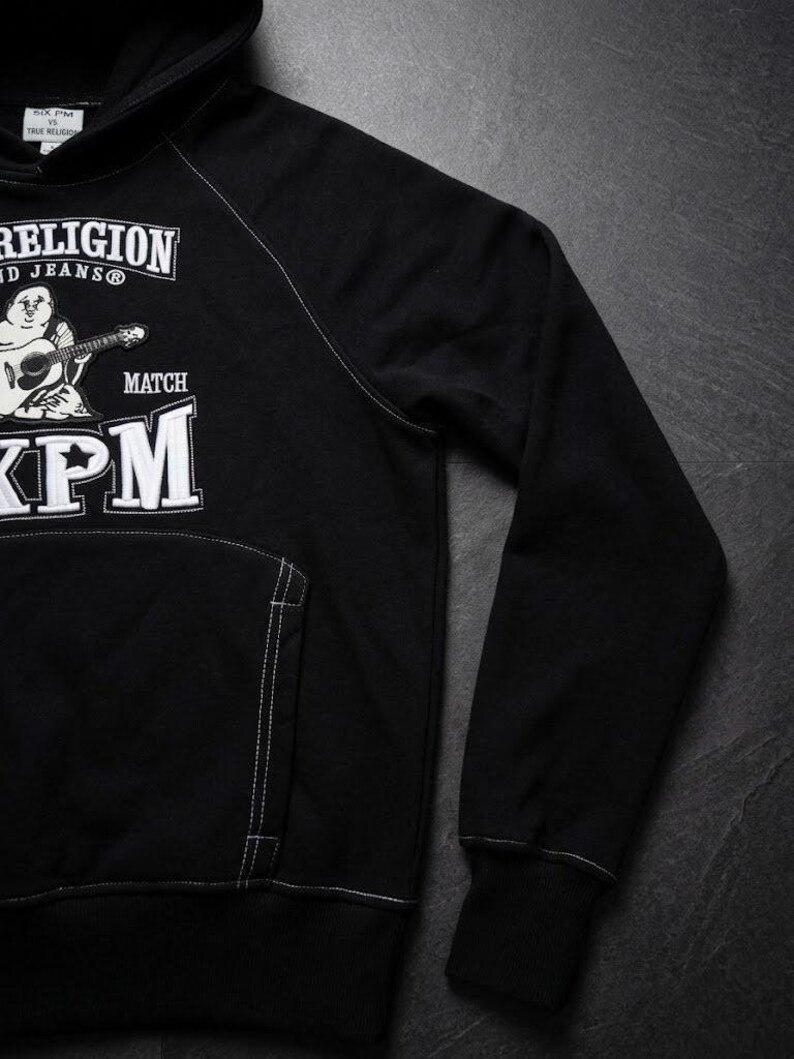 Op de afbeelding: Zwarte hoodie met witte stiksels en een voorzak. De hoodie heeft een afbeelding van een stripfiguur die gitaar speelt, met de tekst "TRUE RELIGION" en "XPM" in het wit.