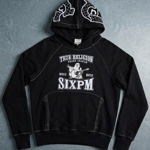 Op de afbeelding: Zwarte hoodie met witte stiksels en een voorzak. De hoodie heeft de witte tekst "TRUE RELIGION BRAND JEANS" en "SIXPM". De capuchon heeft een wit grafisch ontwerp. Een casual, comfortabel kledingstuk.