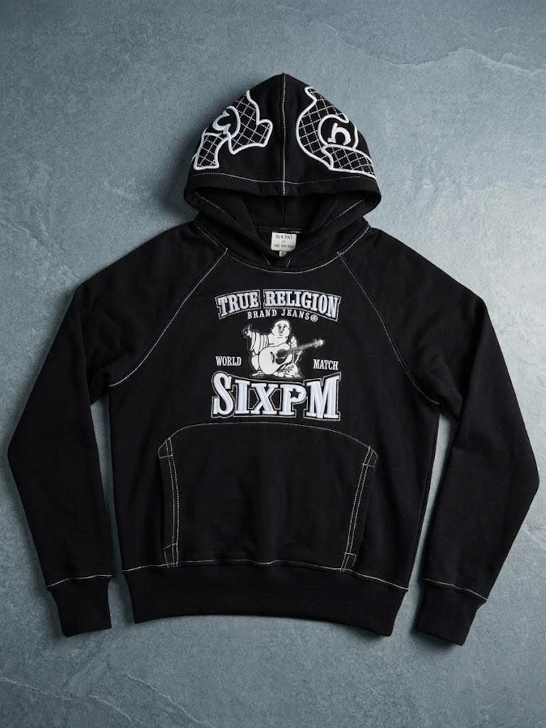 Op de afbeelding: Zwarte hoodie met witte stiksels en een voorzak. De hoodie heeft de witte tekst "TRUE RELIGION BRAND JEANS" en "SIXPM". De capuchon heeft een wit grafisch ontwerp. Een casual, comfortabel kledingstuk.