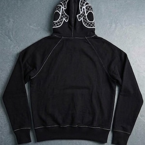 Op de afbeelding: Zwarte hoodie met een wit grafisch ontwerp op de capuchon. De hoodie heeft witte stiksels langs de naden en een kangoeroezak. Het capuchonontwerp is een gestileerd, geometrisch patroon.