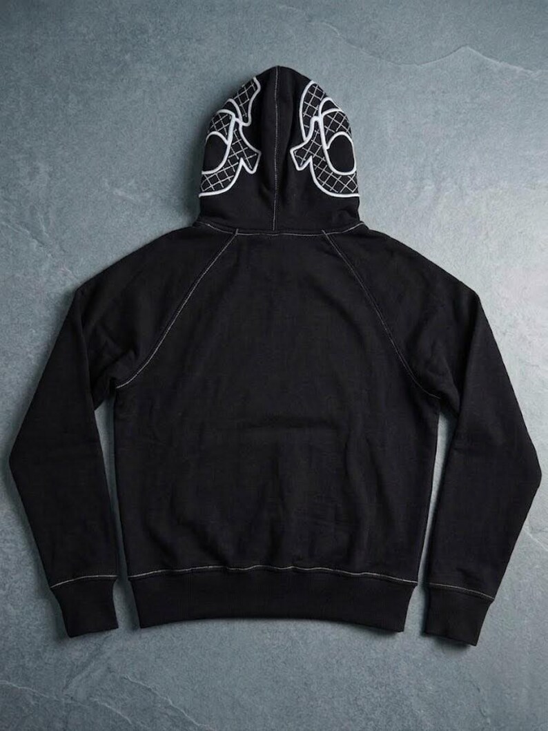 Op de afbeelding: Zwarte hoodie met een wit grafisch ontwerp op de capuchon. De hoodie heeft witte stiksels langs de naden en een kangoeroezak. Het capuchonontwerp is een gestileerd, geometrisch patroon.