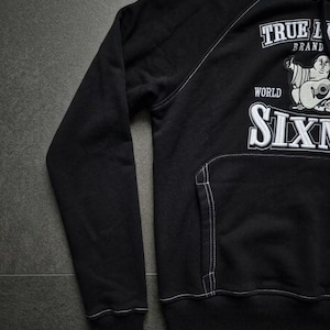 Op de afbeelding: Zwarte hoodie met witte stiksels. Voorzien van een voorzak en een capuchon. De voorkant toont de tekst "TRUE L BRAND" en "WORLD SIXN" met een afbeelding van een cartoon Boeddha die gitaar speelt.