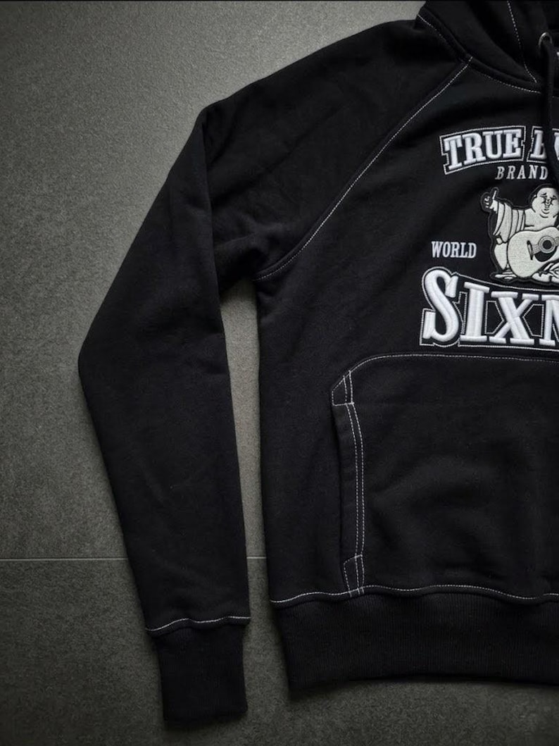 Op de afbeelding: Zwarte hoodie met witte stiksels. Voorzien van een voorzak en een capuchon. De voorkant toont de tekst "TRUE L BRAND" en "WORLD SIXN" met een afbeelding van een cartoon Boeddha die gitaar speelt.