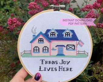 May include: Cross-stitch embroidery in a wooden hoop showing a pink house with a blue roof and the text "Trans Joy Lives Here." The design includes a small fence and a flag. The words "Instant Download PDF Pattern" are also visible.