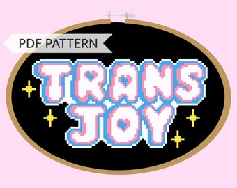 May include: A black embroidery hoop with the words "TRANS JOY" in pixelated letters. The letters are pink, blue, and white with yellow stars around them.