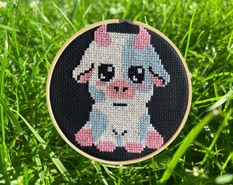 May include: A cross-stitch embroidery hoop with a black background and a white cow with blue and pink accents. The cow has large eyes and a pink nose.