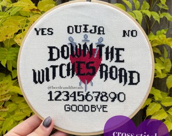 May include: A white cross stitch pattern with a black and red heart and the text "Ouija Down the Witches Road" and "1234567890 Goodbye".