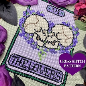 Puede incluir: Patrón de punto de cruz para una carta de tarot, Los Enamorados, con dos calaveras blancas con flores moradas. La carta está etiquetada con el número romano VI y la palabra "Enamorados".