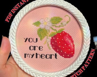 May include: A pink cross stitch pattern with a white frame. The pattern features a red strawberry with green leaves and a white flower. The text "You are my heart" is stitched in black.