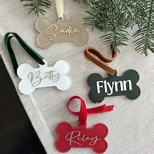 May include: Four bone-shaped Christmas ornaments with different colored backgrounds and gold lettering. The ornaments are labeled "Sadie", "Betty", "Flynn", and "Riley".