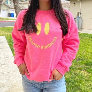 Pode incluir: Uma camisa de moletom rosa com um rosto sorridente amarelo e o texto "choose kindness" impresso na frente.