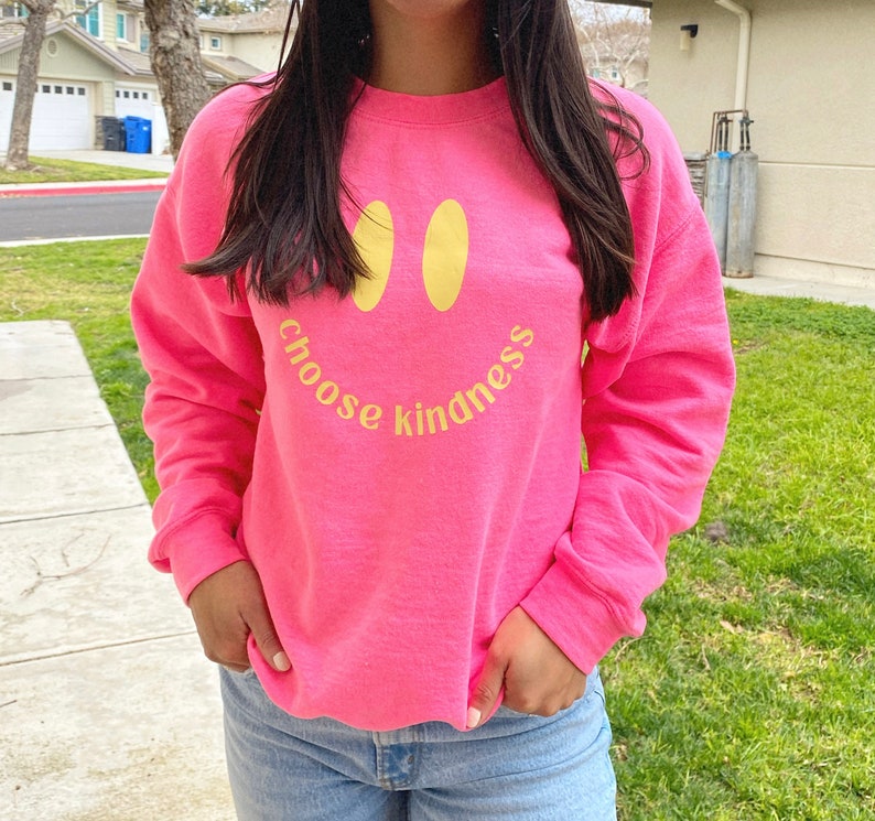 Pode incluir: Uma camisa de moletom rosa com um rosto sorridente amarelo e o texto "choose kindness" impresso na frente.
