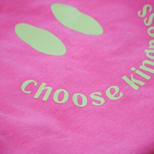 Pode incluir: Camiseta rosa com um rosto sorridente amarelo e o texto "choose kindness" impresso nela.