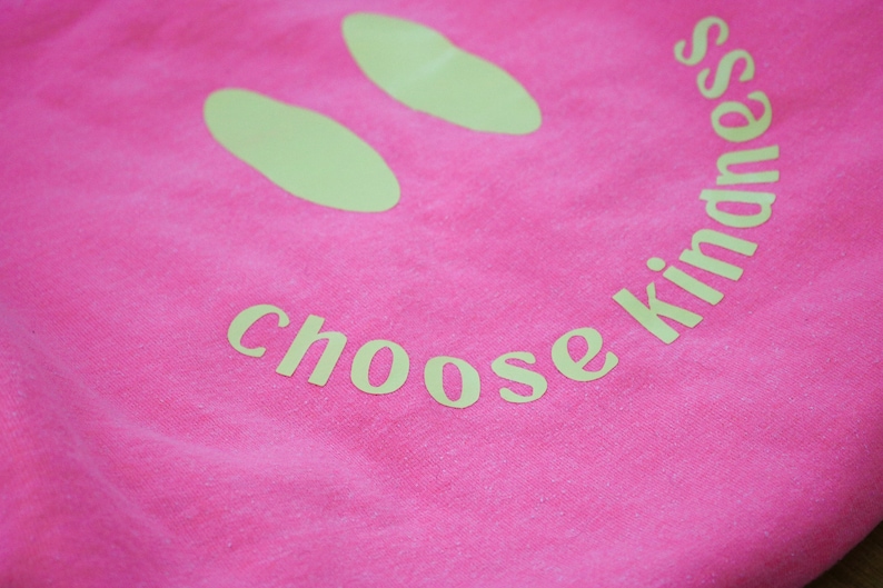 Pode incluir: Camiseta rosa com um rosto sorridente amarelo e o texto "choose kindness" impresso nela.
