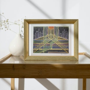 May include: A framed print of a colorful abstract geometric design with a cityscape in the background. The print is in a gold frame and is sitting on a wooden table.