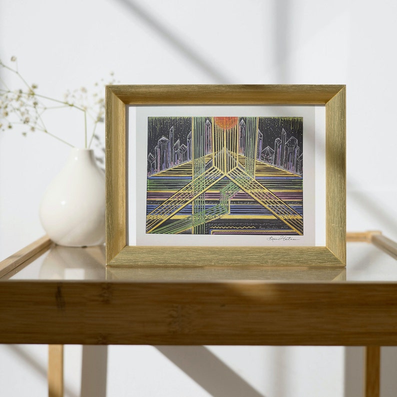 May include: A framed print of a colorful abstract geometric design with a cityscape in the background. The print is in a gold frame and is sitting on a wooden table.