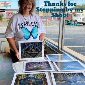 May include: A woman wearing a white t-shirt with a blue butterfly graphic and the word "Fearless" printed on it, is holding a framed print of a geometric design.  She is standing in front of a window with a sign that says "Thanks for stopping by my shop! Becca".  There are five framed prints on a table in front of her, each with a different colorful design.