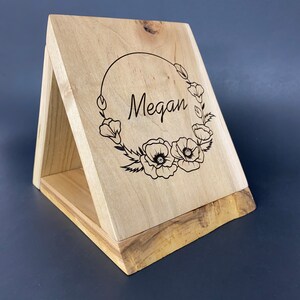 Puede incluir: Una caja de madera con tapa abatible, que presenta un diseño grabado con láser de una corona floral con el nombre "Megan" en su interior. La caja está hecha de madera clara con un acabado natural.