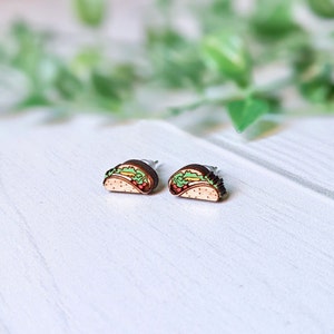 May include: A pair of stud earrings shaped like tacos. Each earring is crafted from wood and features a detailed, colorful illustration of a taco with a light tan shell, green lettuce, and red tomato accents, finished with a metal post backing.