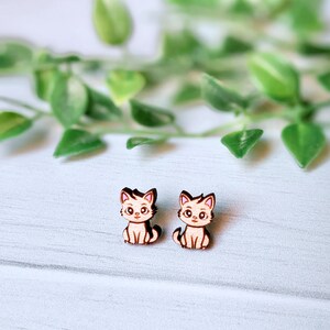 Pode incluir: Um par de brincos de gato de madeira. Os gatos são de cor castanho claro com narizes cor-de-rosa e olhos brancos. Os brincos são pequenos e delicados.