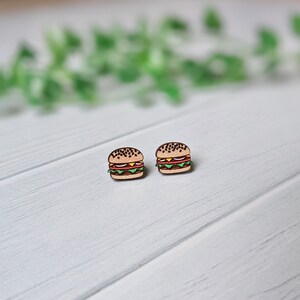 May include: A pair of wooden earrings shaped like hamburgers. The earrings are brown with black sesame seeds on the top bun, a yellow cheese slice, a red tomato slice, and a green lettuce leaf.