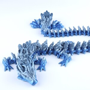 May include: A 3D printed articulated dragon figurine featuring a metallic blue and silver gradient finish. The creature has a detailed head with horn-like protrusions, four clawed feet, and a long, segmented body that curves across the frame.
