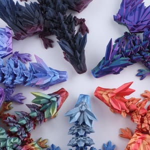Puede incluir: Una colecci&oacute;n de cinco figuras de drag&oacute;n articuladas con una textura facetada similar al cristal. Los dragones est&aacute;n coloreados en degradados de morado oscuro, azul y morado iridiscente, azul claro, verde y amarillo, y naranja y rojo.