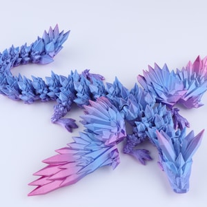 Puede incluir: Una figura de drag&oacute;n articulada con una textura geom&eacute;trica cristalina. El cuerpo presenta un degradado de color azul profundo a magenta vibrante, con p&uacute;as angulares afiladas que cubren la cabeza, las alas, el lomo y la larga cola curvada.