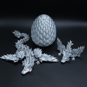 Puede incluir: Un conjunto impreso en 3D de color plateado que consta de un huevo grande y texturizado con un patr&oacute;n de escamas geom&eacute;tricas, una figura de drag&oacute;n larga y articulada con segmentos cristalinos afilados y un drag&oacute;n m&aacute;s peque&ntilde;o a juego con las alas extendidas.