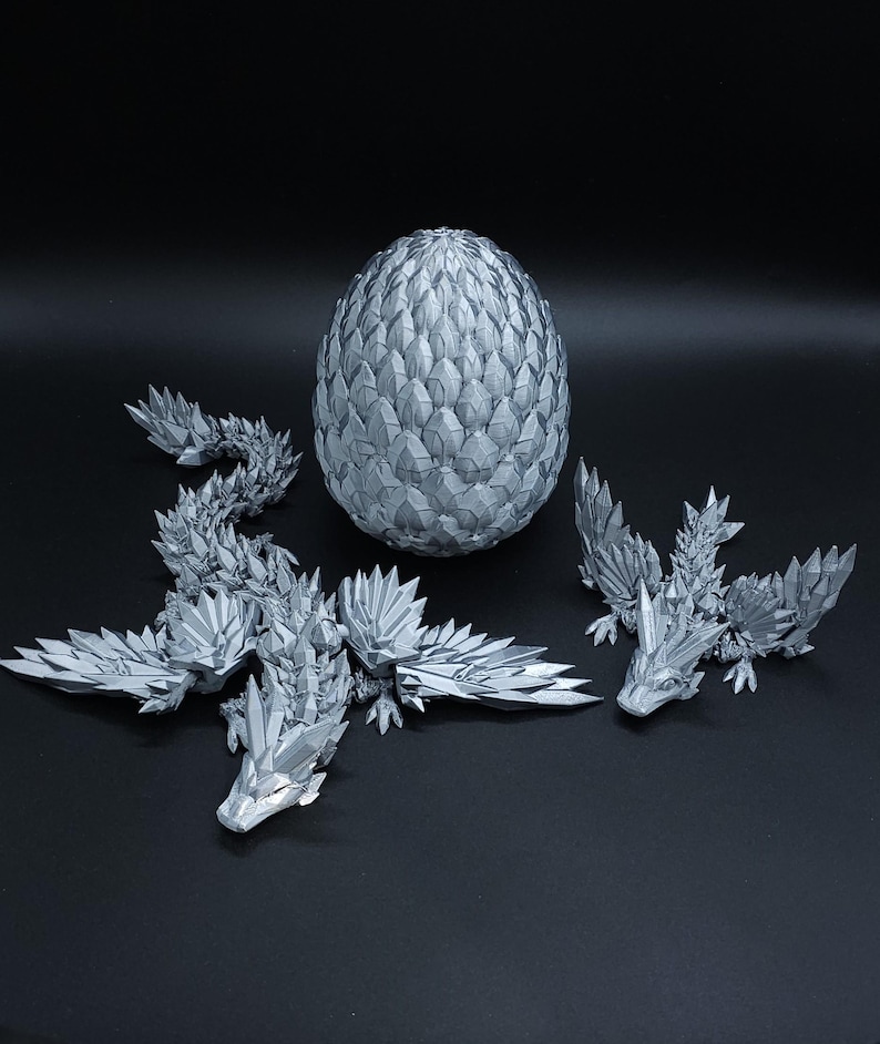 Puede incluir: Un conjunto impreso en 3D de color plateado que consta de un huevo grande y texturizado con un patr&oacute;n de escamas geom&eacute;tricas, una figura de drag&oacute;n larga y articulada con segmentos cristalinos afilados y un drag&oacute;n m&aacute;s peque&ntilde;o a juego con las alas extendidas.