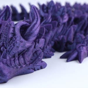 May include: A 3D printed articulated dragon figure in a deep metallic purple finish. The creature features a detailed head with prominent horns, a snout, and a segmented, flexible body composed of overlapping scales that extend across the frame.