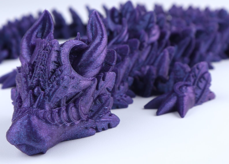 May include: A 3D printed articulated dragon figure in a deep metallic purple finish. The creature features a detailed head with prominent horns, a snout, and a segmented, flexible body composed of overlapping scales that extend across the frame.
