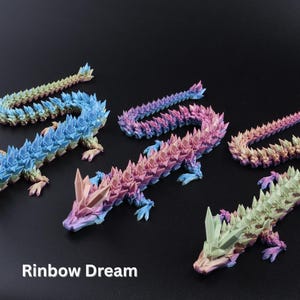 May include: Five colorful dragon figurines in shades of blue, pink, purple, and green. Each dragon has a spiky, crystal-like design and is posed on a dark surface. The text "Rainbow Dream" is at the bottom.