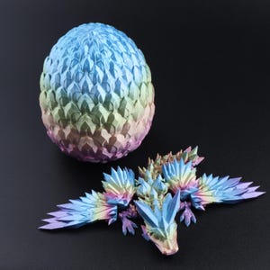 Puede incluir: Una figura de drag&oacute;n articulada impresa en 3D con un acabado degradado multicolor en azul, verde, amarillo y morado, con escamas cristalinas afiladas. A su lado hay un recipiente con forma de huevo a juego con la misma textura de escamas.