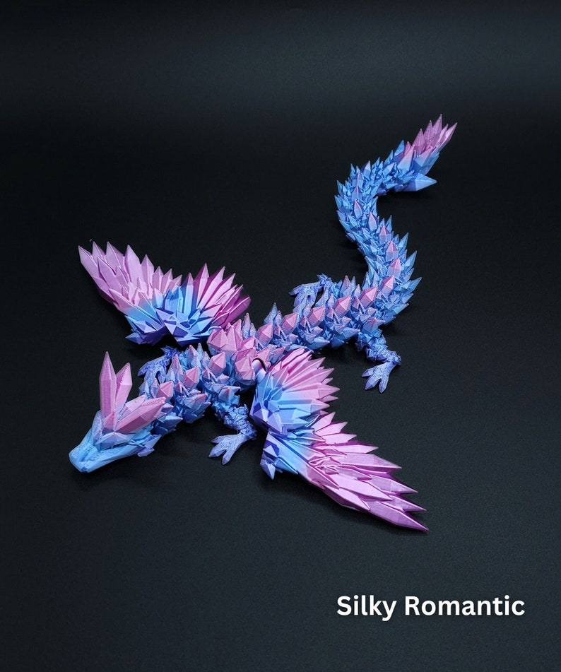 Puede incluir: Una figura de drag&oacute;n articulada impresa en 3D con un degradado de colores azul y rosa, con p&uacute;as afiladas similares a cristales que cubren su cuerpo, alas y cabeza. El texto Silky Romantic est&aacute; impreso en blanco en la esquina inferior derecha.