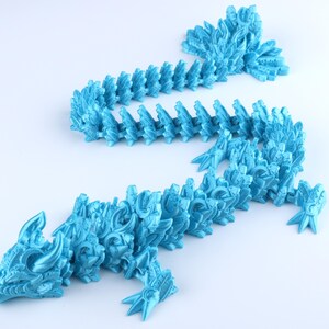 May include: A bright cyan articulated dragon figurine featuring a segmented body with intricate scale patterns, a detailed head with horns, and a long tail. The flexible design allows the creature to be posed in various positions on a flat surface.