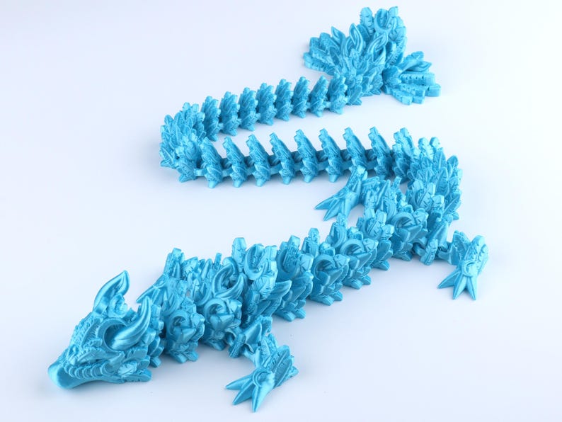 May include: A bright cyan articulated dragon figurine featuring a segmented body with intricate scale patterns, a detailed head with horns, and a long tail. The flexible design allows the creature to be posed in various positions on a flat surface.